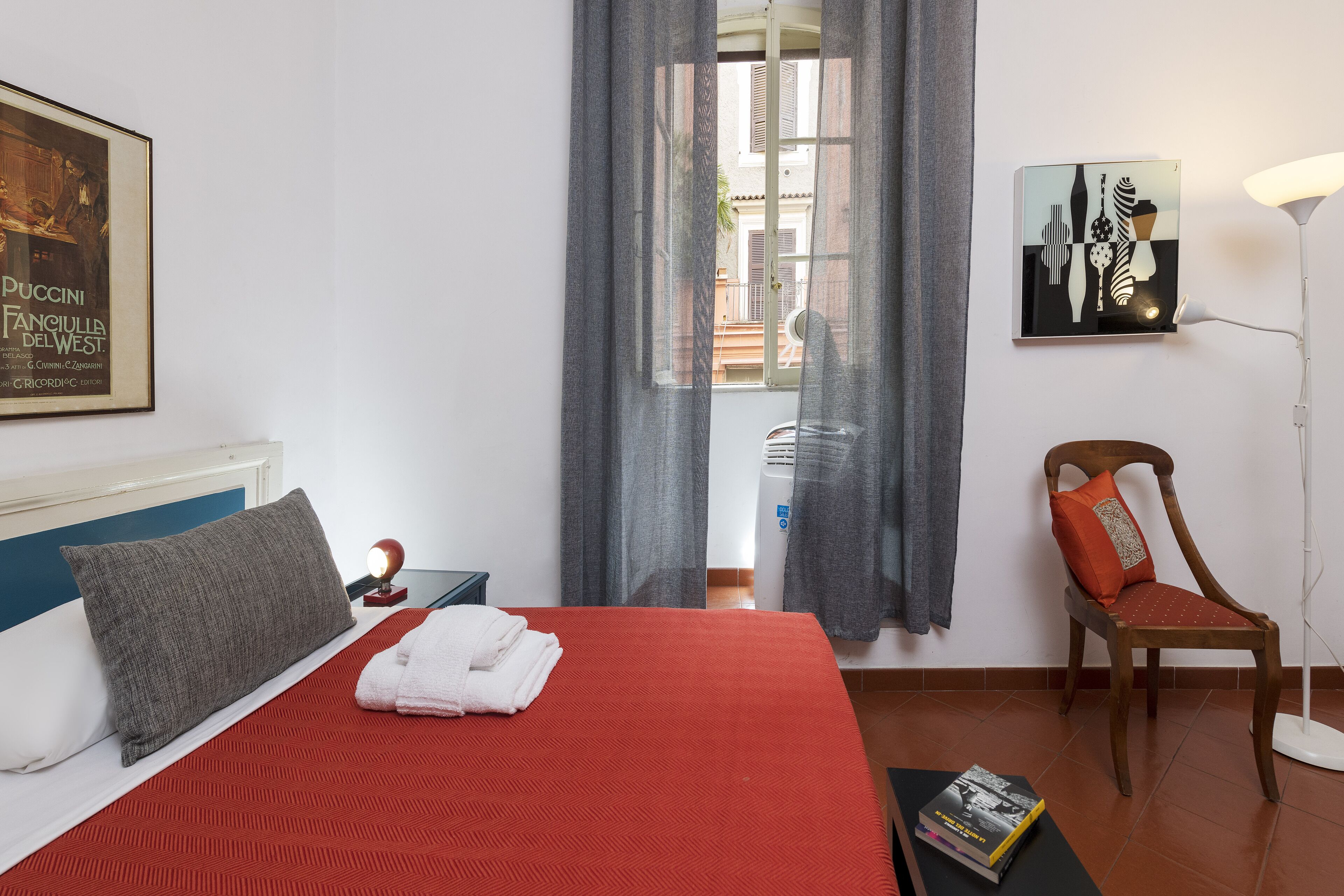 A neatly made bed with a red bedspread, a wooden chair with an orange cushion, a framed picture on the wall, and a lamp on a side table.