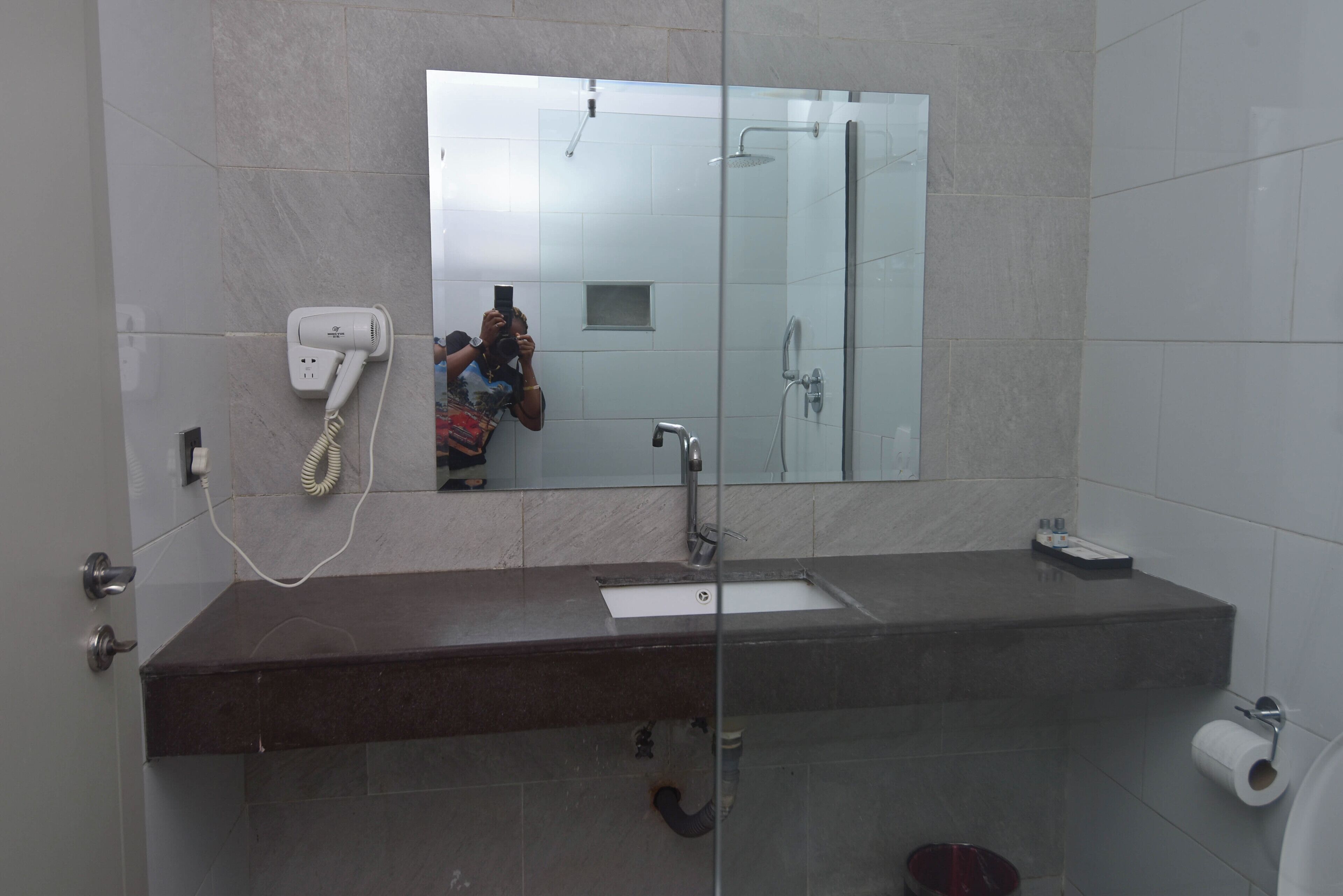 Une salle de bain avec une douche à parois de verre, un lavabo blanc, un miroir et un sèche-cheveux fixé au mur.
