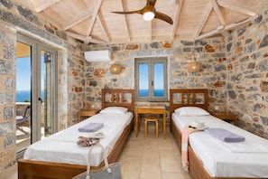 Une pièce avec deux lits en bois, un mur en pierre, un ventilateur de plafond, une fenêtre donnant sur un paysage agréable et une table avec des chaises.