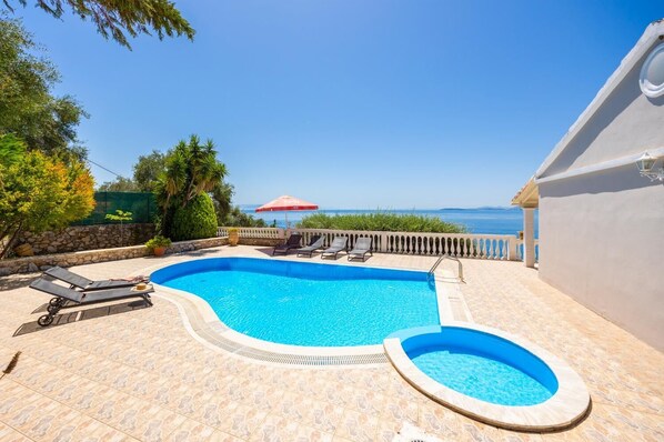 A swimming pool with two sun loungers and a small hot tub, surrounded by a stone patio and a view of the sea.