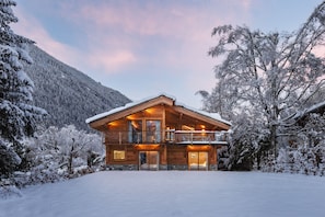 Uma cabana de madeira com varanda, rodeada por árvores cobertas de neve e uma montanha ao fundo.