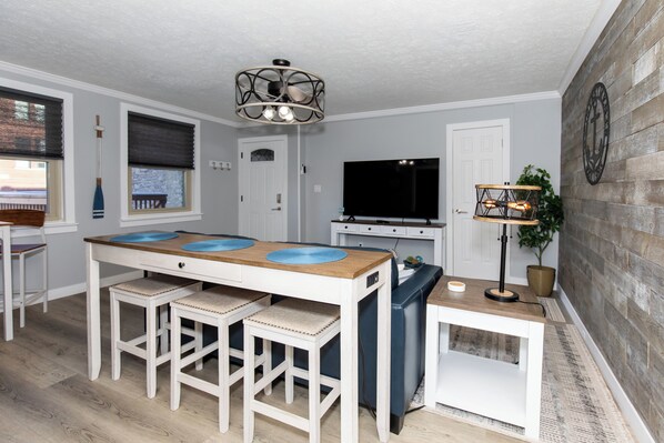A modern kitchen with a wooden table, bar stools, a built-in oven, a television, and a decorative wall clock. - Cozy Downtown Waterfront Escape (Sandusky)