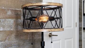 Eine Tischlampe mit Holzfuß und schwarzem Metallgestell auf einem Holztisch vor einer rustikalen Ziegelwand.
