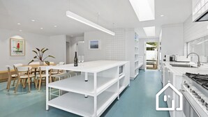 A modern kitchen with white cabinetry, a central island, and a dining area with a table and chairs. - Villa Santorini *1 NIGHT FREE* Secluded Back Beach Poolside Oasis (Blairgowrie)