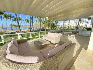 A covered outdoor seating area with wicker furniture and a view of the beach and palm trees.