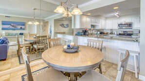 A modern kitchen with white cabinets, a central island, and a dining area with a round table and chairs. - B404 | Beachfront with Beach Service, Pool, & WiFi (Miramar Beach)