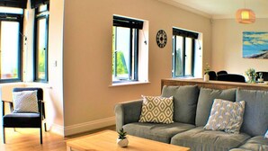 Ein Wohnzimmer mit grauem Sofa, einem Holztisch und einem Sessel mit gemusterten Kissen. Zwei Fenster mit schwarzen Rahmen sowie eine Uhr an der Wand sind zu sehen.