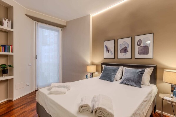 Una cama bien hecha con toallas, una mesita de noche con lámpara, y una estantería con libros y una planta.