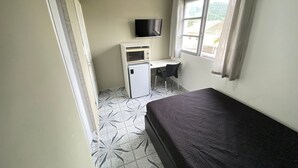 A small room with a bed, a desk, a television, and a refrigerator. - Suites - Foot on the sand Canto do Forte Beach (Praia Grande)