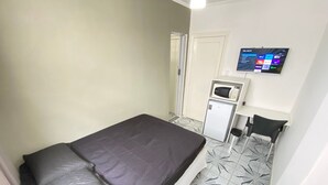Una habitación pequeña y funcional con cama, microondas, refrigerador y televisor en la pared.