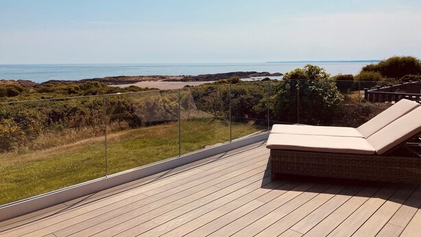 Une terrasse en bois avec un fauteuil de relaxation et une balustrade en verre donnant sur un paysage côtier.