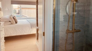 Une chambre à coucher avec un lit, des oreillers et une table de chevet. On y trouve une cabine de douche en verre avec un pommeau de douche et un flexible dorés.