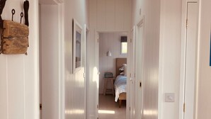 Un couloir étroit aux murs blancs et au plancher en bois, qui mène à une chambre à coucher avec un lit et une fenêtre.