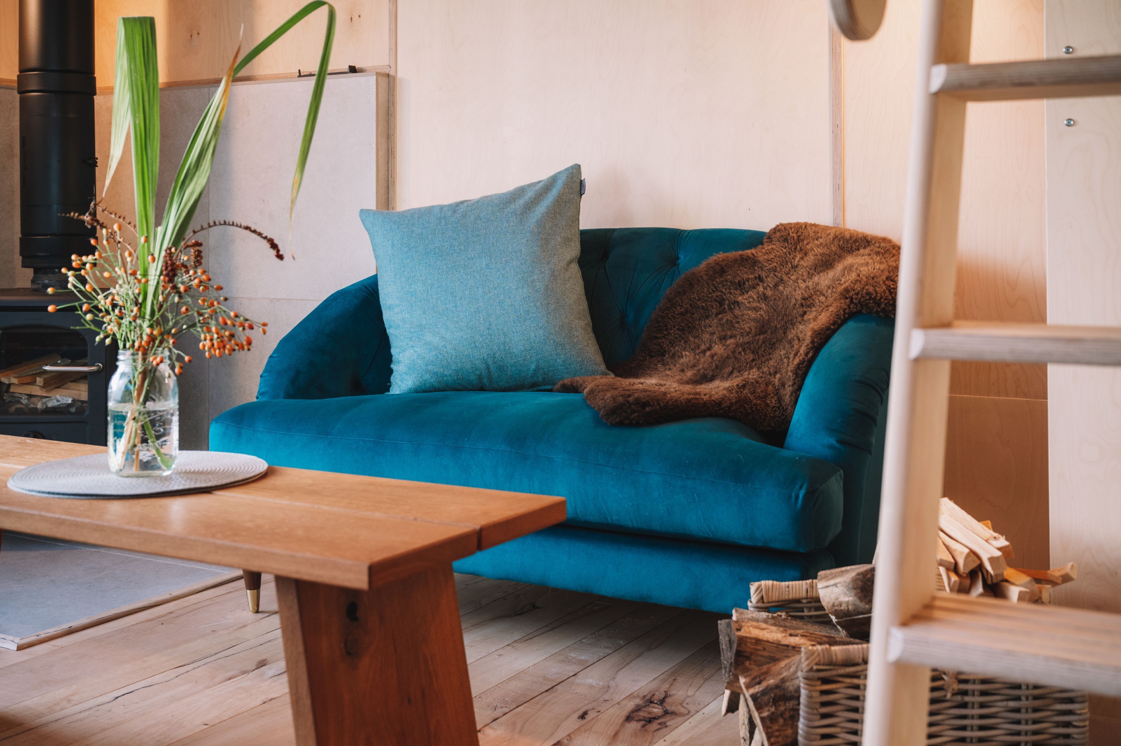 Un canapé de couleur bleu sarcelle agrémenté d’un coussin bleu et d’un plaid marron, une table basse en bois et un vase contenant des plantes séchées.
