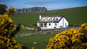 A white house with a sloped roof, large windows, and a stone wall in the foreground.