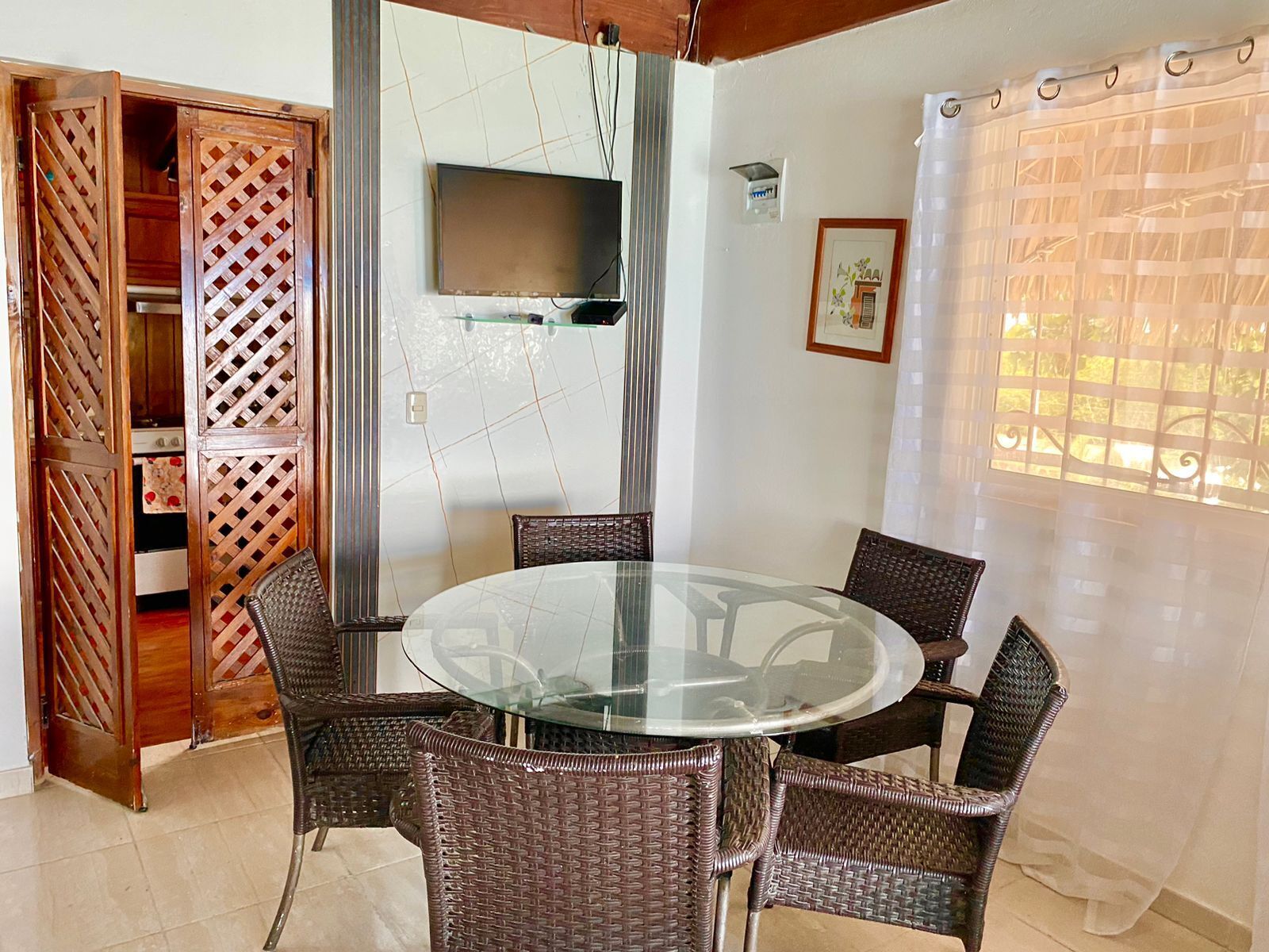 A dining area with a round glass table surrounded by wicker chairs, a TV mounted on the wall, and a window with sheer curtains.