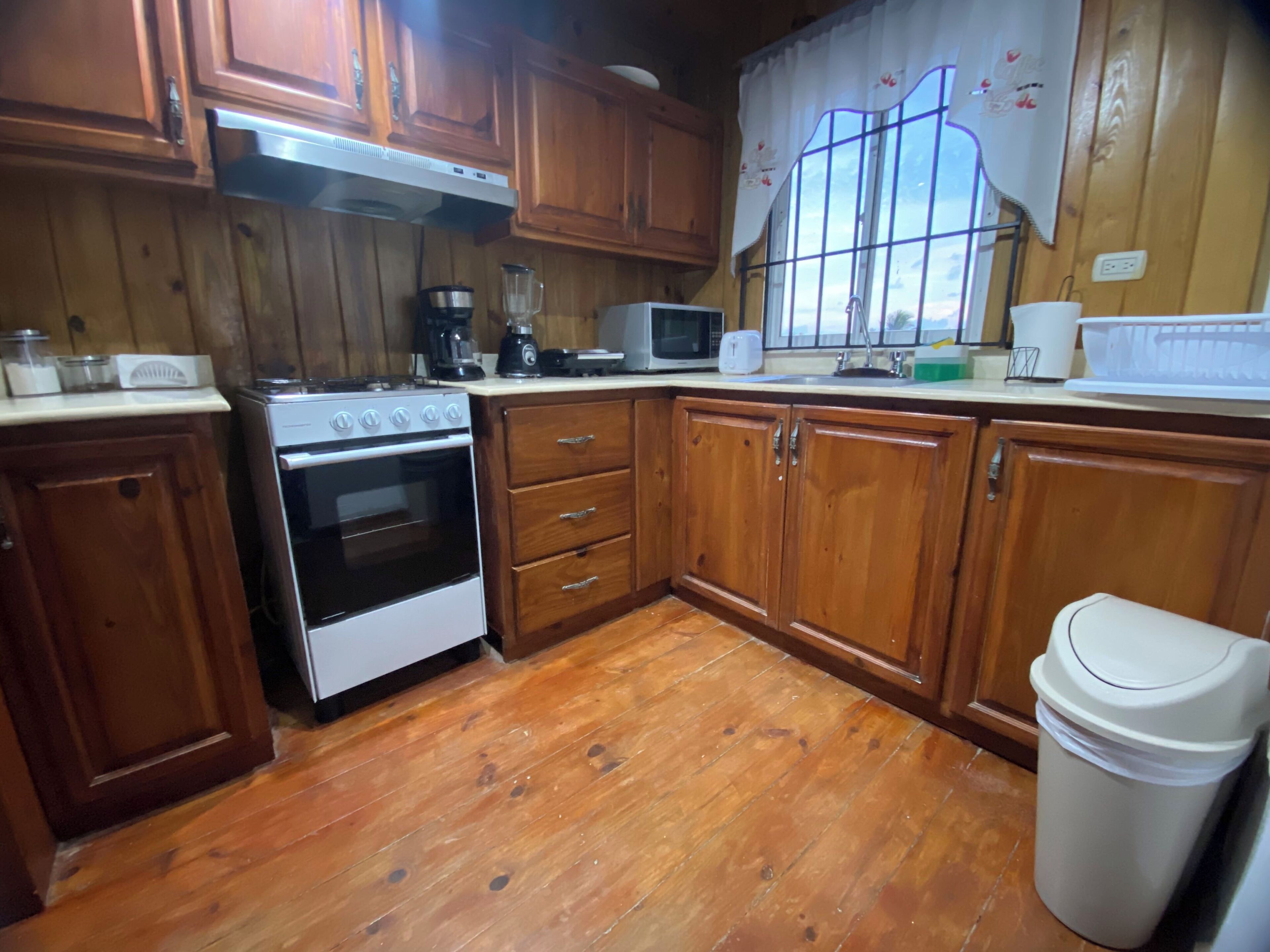 A kitchen with wooden cabinets, a stove, a microwave, and a trash can.