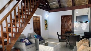 A spacious living area with a wooden staircase, wicker furniture, a dining table, and a television. - Hostal Magallanes Bayahibe (San Rafael del Yuma)