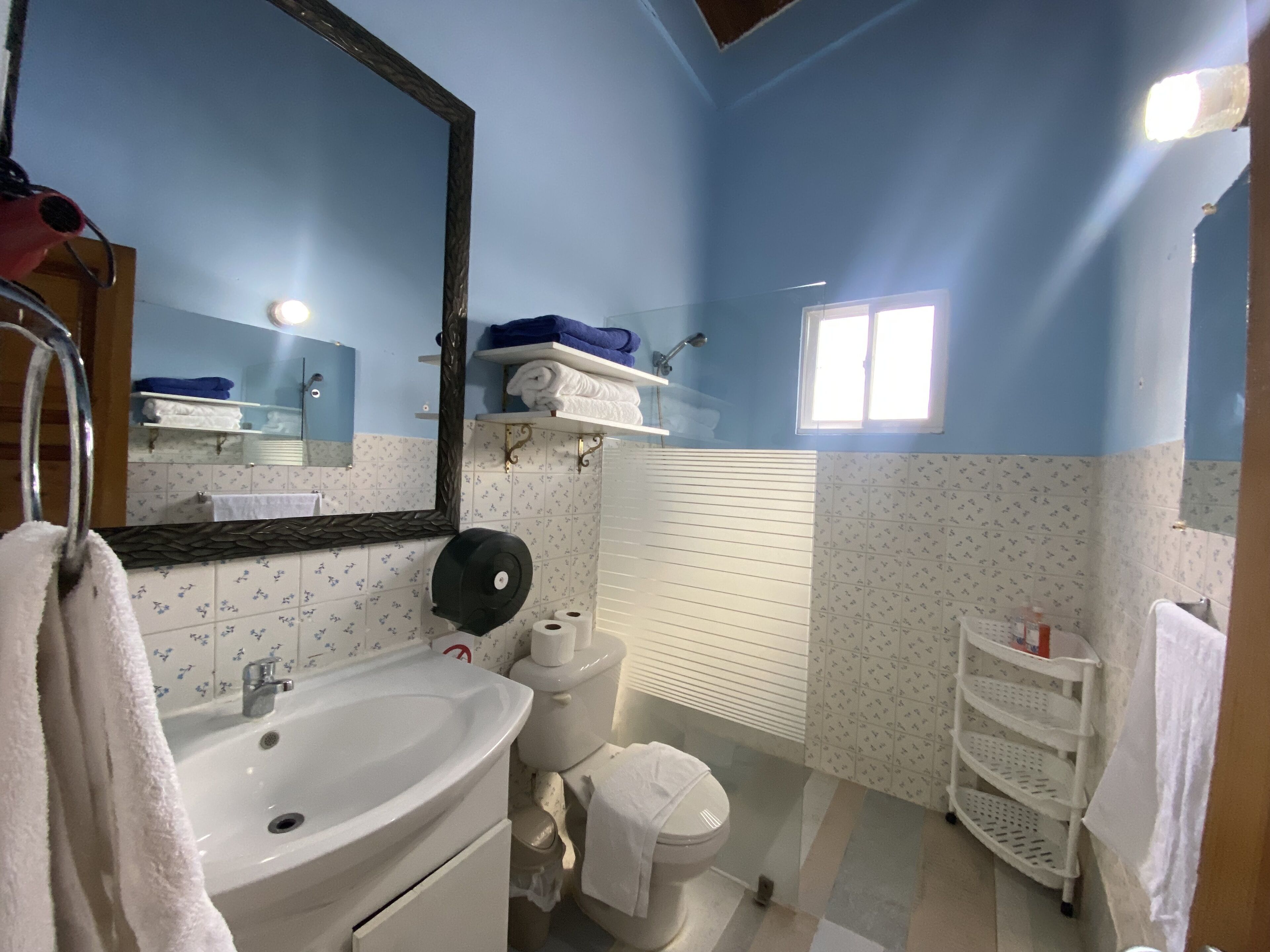 Baño con lavamanos, inodoro y espejo. Hay toallas y un estante con artículos de aseo.