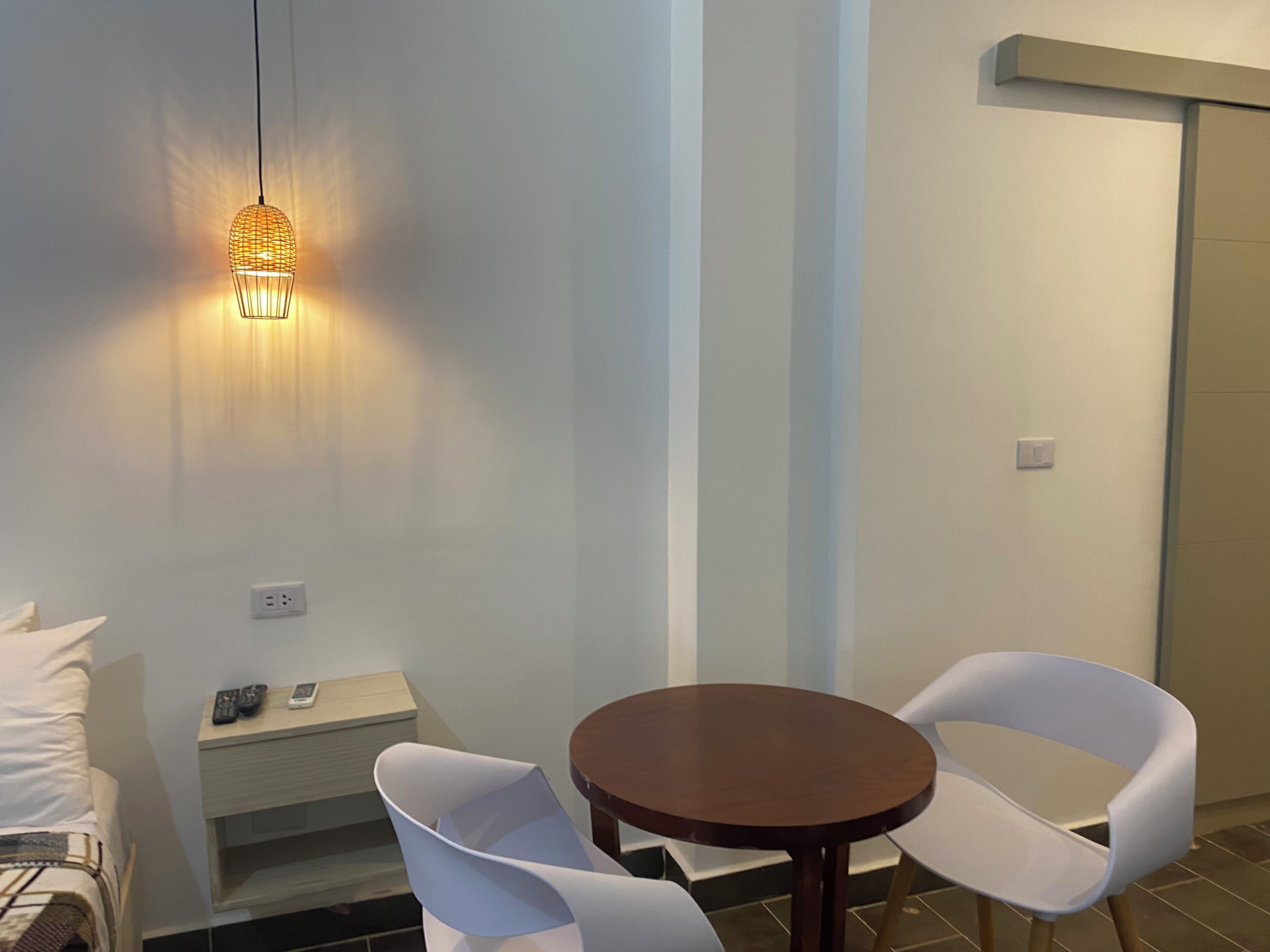 A hotel room with a bed, bedside table with a phone, a small round table with two white chairs, and a hanging light fixture.