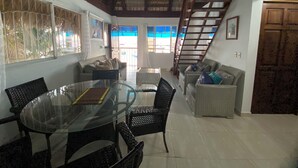 A spacious living area with a glass dining table surrounded by wicker chairs, a sofa, and a staircase leading to an upper level. - Hostal Magallanes Bayahibe (San Rafael del Yuma)