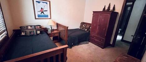 A bedroom with two beds, a wooden wardrobe, a framed picture on the wall, and a small table.