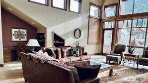 A spacious living room with a large brown sofa, a coffee table, and a fireplace. There are multiple windows providing natural light and a view of the outside. - Ski-In/Out 5 BR House at Copper Mtn (Frisco)