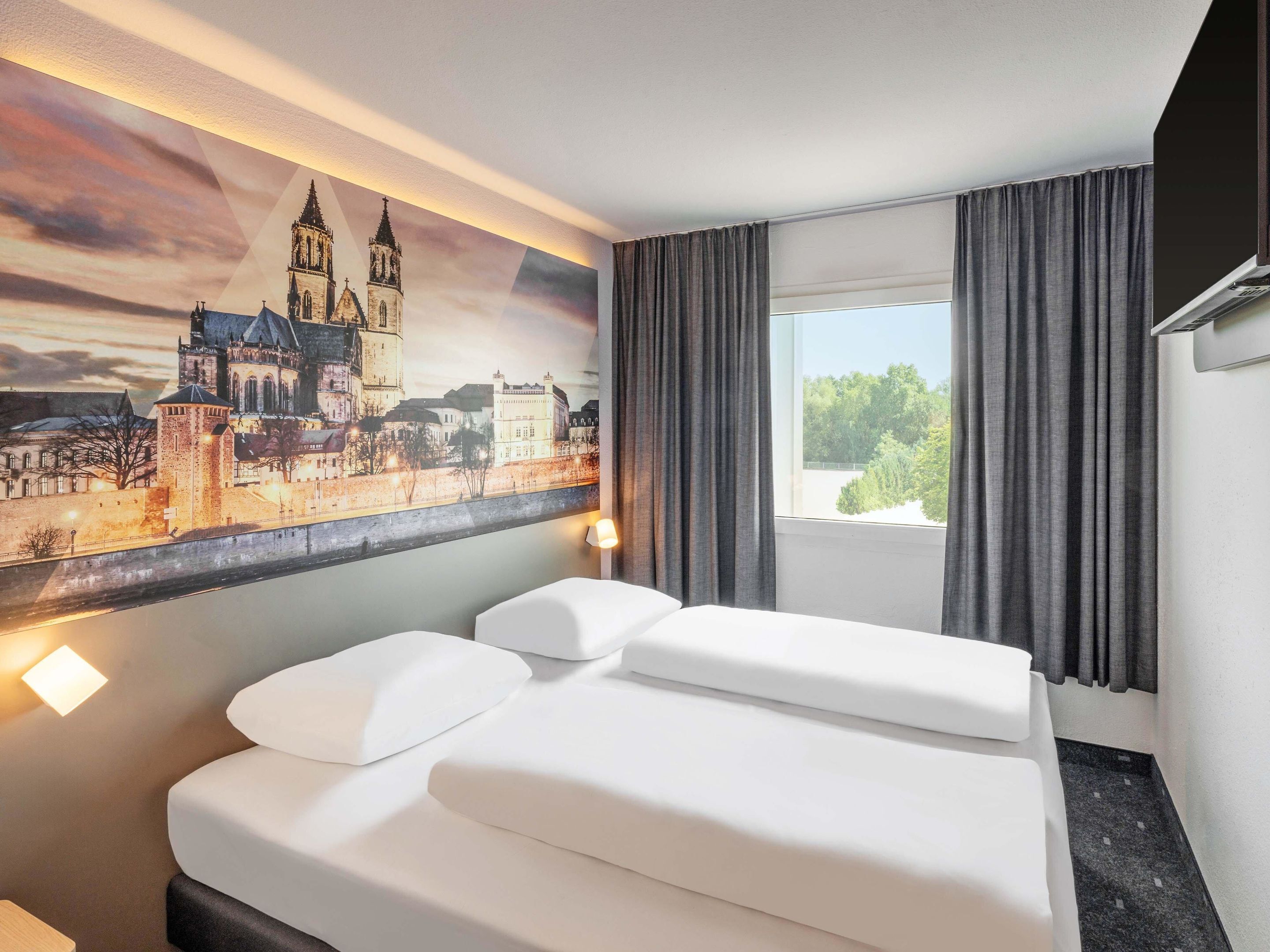 A hotel room with a bed, a large window with curtains, and a cityscape painting on the wall.
