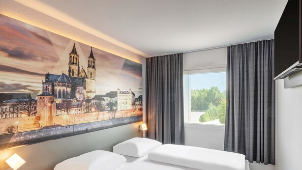 A hotel room with a bed, a large window with curtains, and a cityscape painting on the wall. - B&B HOTEL Magdeburg-Barleben (Barleben)