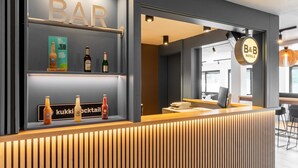 Un espace bar moderne avec un comptoir d’accueil en bois, des étagères où sont exposées des bouteilles et une enseigne indiquant le nom du bar.