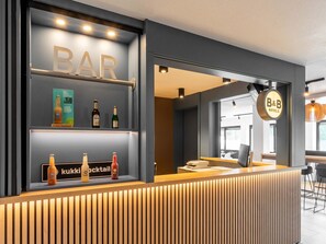 Un espace bar moderne avec un comptoir d’accueil en bois, des étagères où sont exposées des bouteilles et une enseigne indiquant le nom du bar.