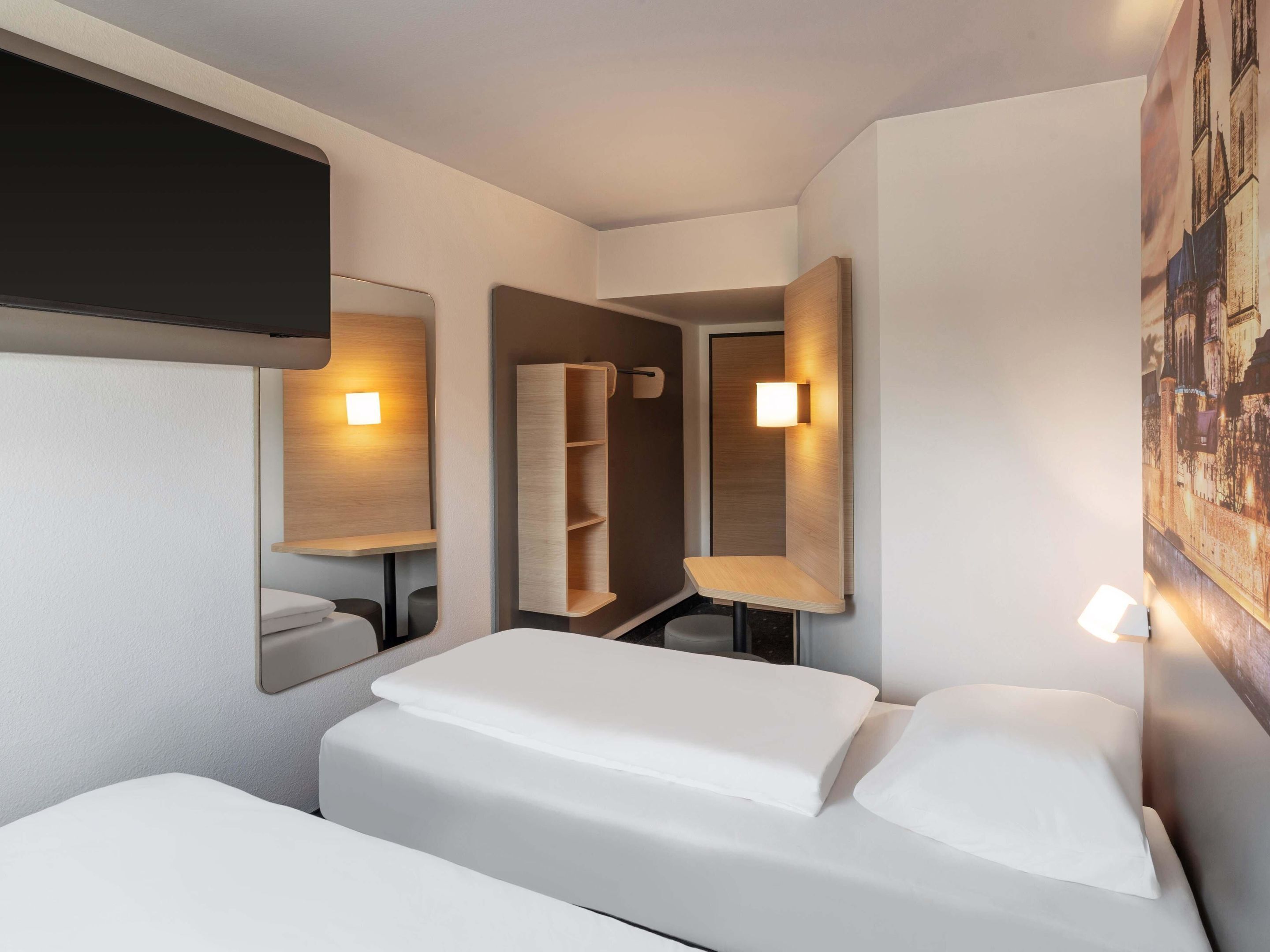 A hotel room with two beds, a mirror, a shelf, and a wall-mounted light.