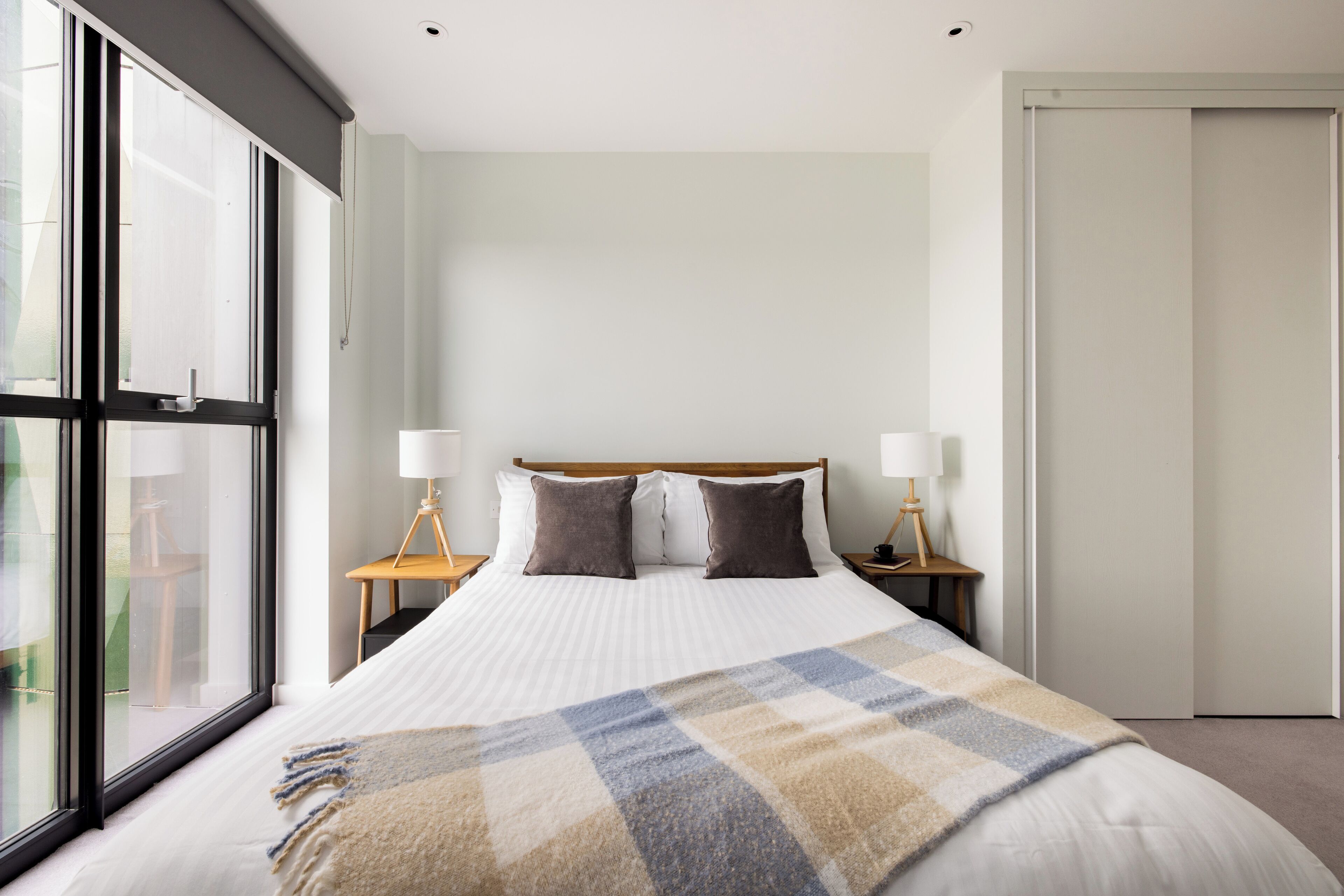 A modern hotel room with a large bed, two bedside tables with lamps, a view through floor-to-ceiling windows, and a closed door.