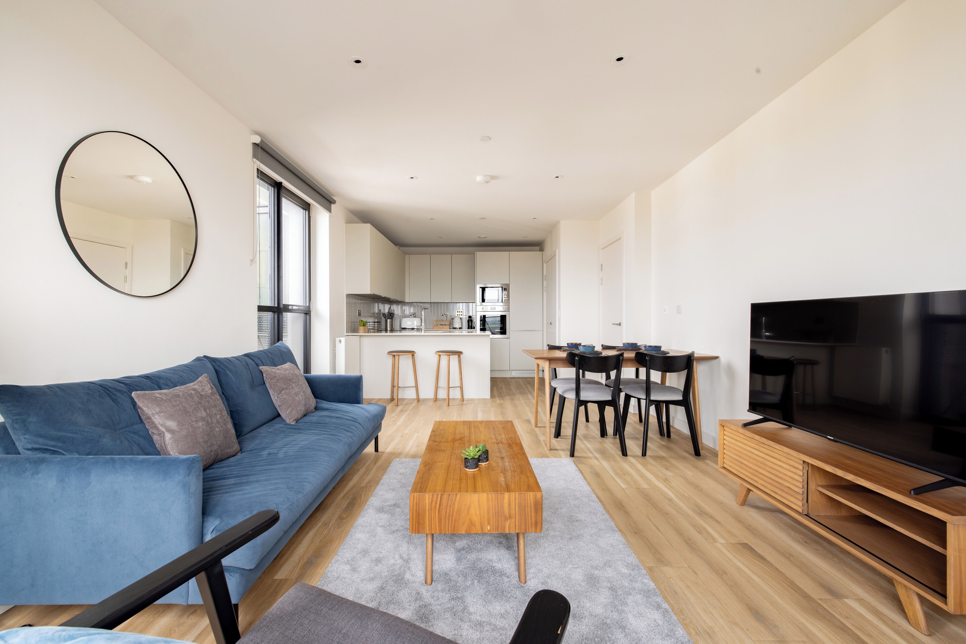Een moderne woonkamer met een blauwe bank, een houten salontafel, een televisie en een keuken op de achtergrond.