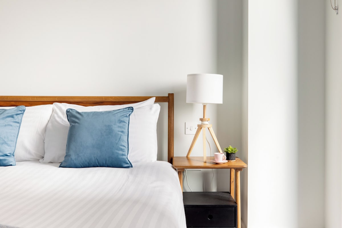 A neatly made bed with blue and white pillows, a bedside table with a lamp, and a small potted plant.