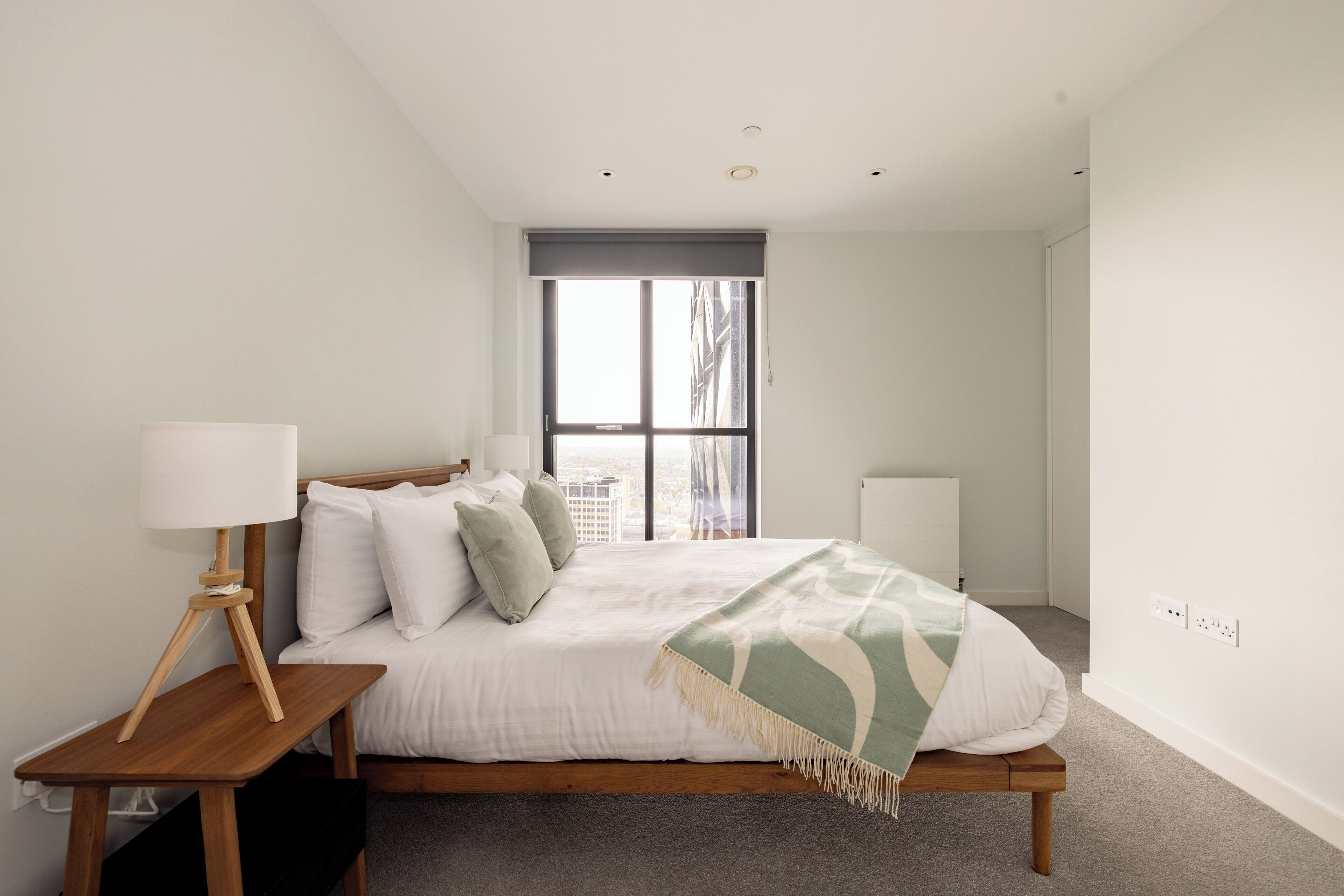 Une chambre à coucher avec un lit, une table de chevet, une lampe et une fenêtre donnant sur des immeubles.