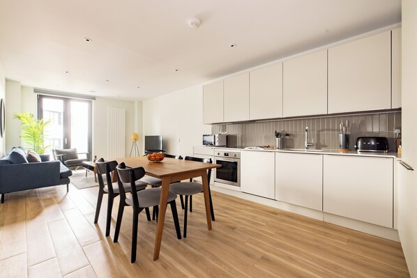 A modern kitchen with white cabinets, a dining area with a wooden table and chairs, and a living room with a blue sofa. - Croydon by Charles Hope (Croydon)