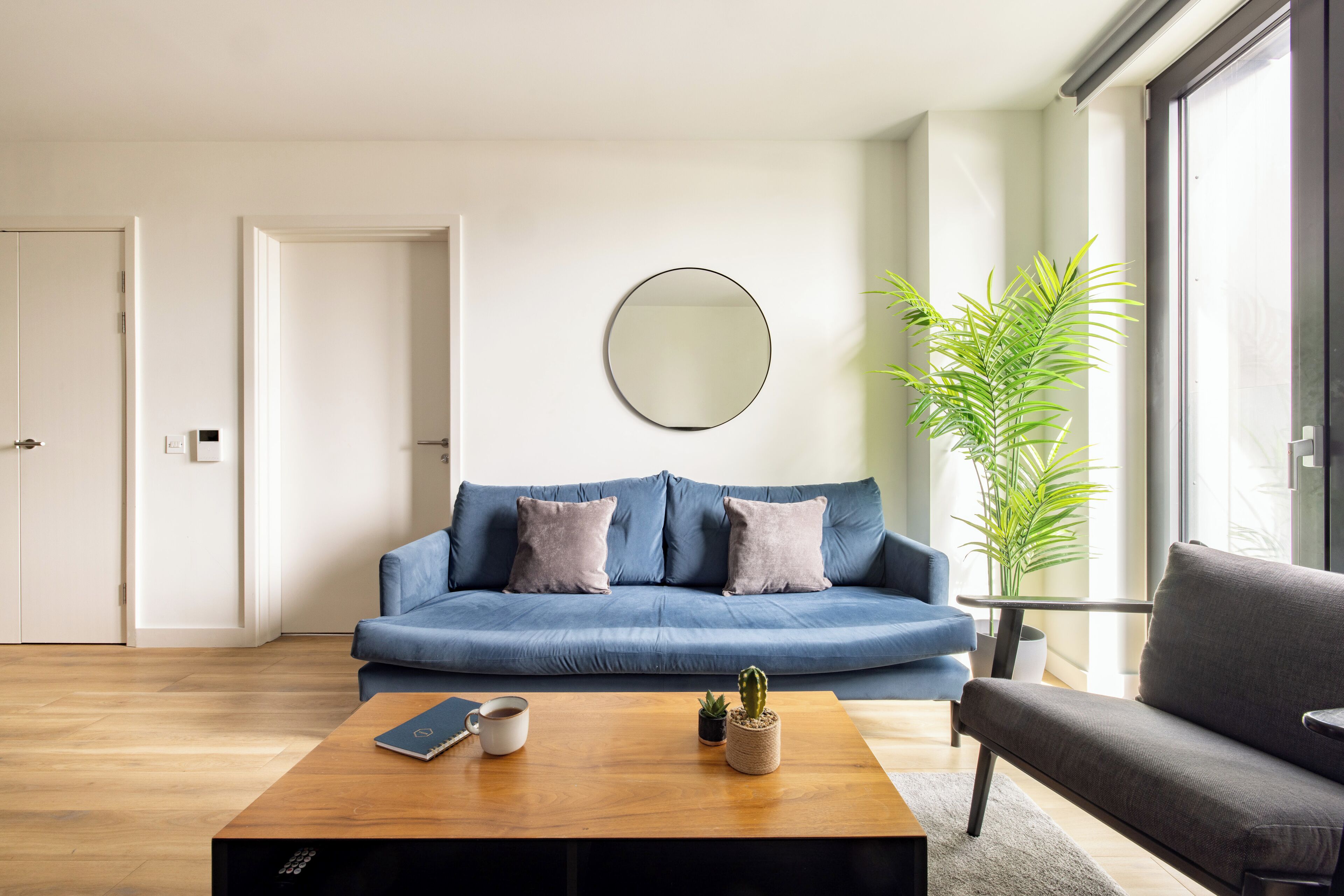 A modern living room with a blue sofa, a round mirror, a coffee table with a book and a mug, and a potted plant.
