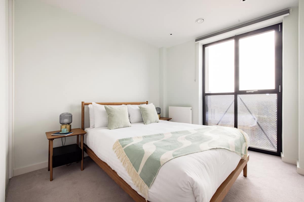 A bedroom with a bed, bedside table, lamp, and a large window with a view.