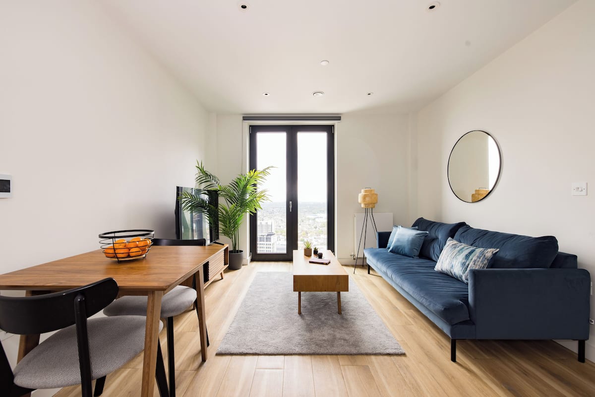 A modern living room with a wooden dining table, a blue sofa, and a large window with a city view.