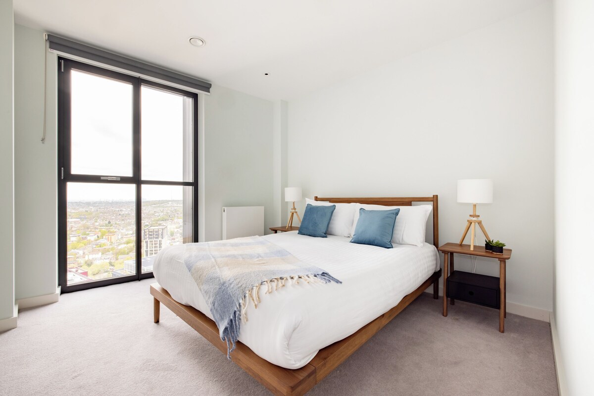 A modern bedroom with a large bed, two bedside tables with lamps, a view through a sliding door, and a cityscape outside.