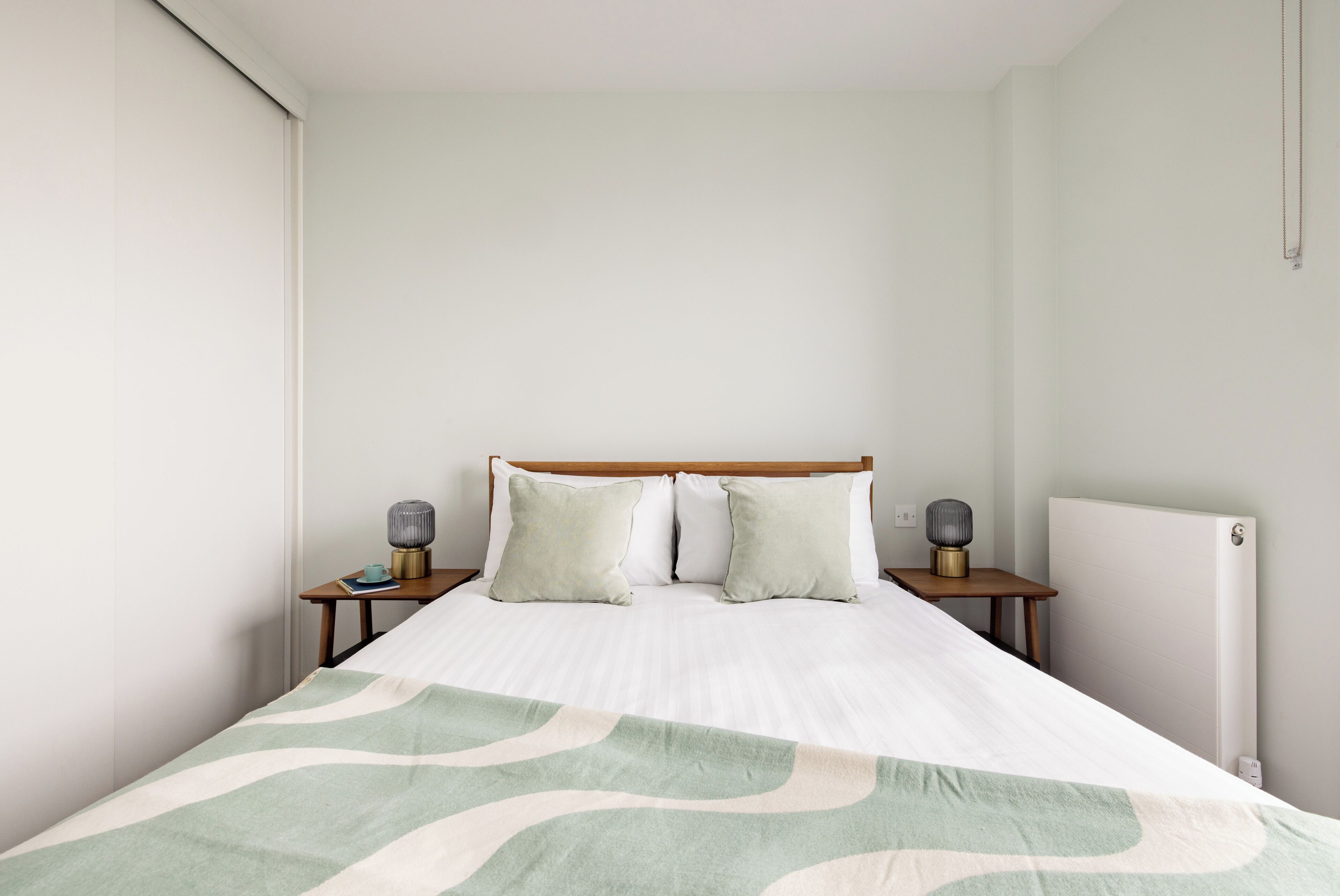 Een net opgemaakt bed met een beddekleed met patroon, twee nachtkastjes met lampen en een radiator aan de muur.