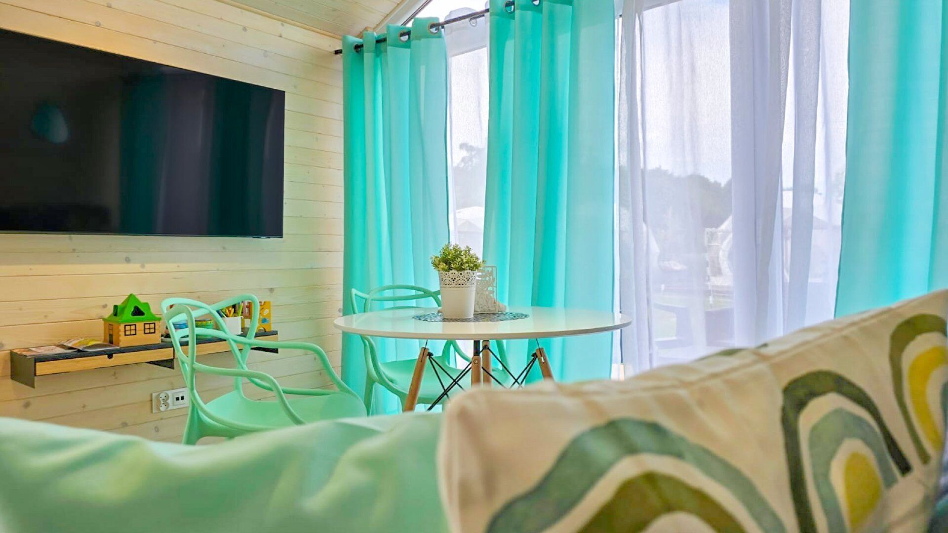 Una habitación acogedora con televisión montada en la pared, una mesa redonda con sillas y una ventana con cortinas color turquesa.