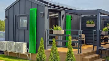 Un edificio moderno e compatto, con facciata grigia, una zona salotto esterna coperta e un piccolo giardino con piante in vaso.