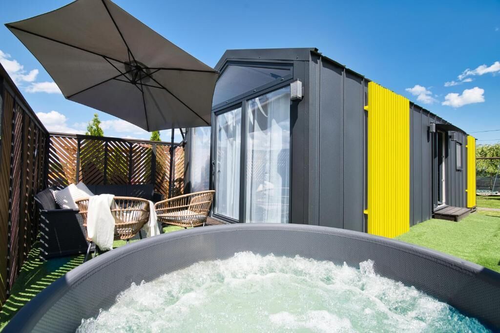A hot tub with a wooden deck and wicker chairs, a covered patio area, and a small building with a yellow accent wall.