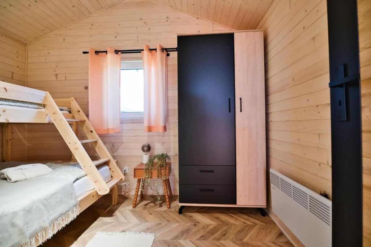 Ein gemütliches Blockhauszimmer mit einem Etagenbett, einem Kleiderschrank, einem kleinen Tisch mit Pflanze und einem Fenster mit Vorhängen.