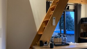 Een houten trap met een ingebouwde werkplek en kasten, een raam met uitzicht op bomen, en een keukenhoek met een magnetron en waterkoker.