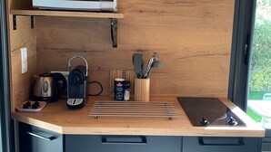 Een kleine keuken met een magnetron, waterkoker en broodrooster op een houten aanrecht. Daaronder zijn kastjes en een raam met uitzicht op groen.
