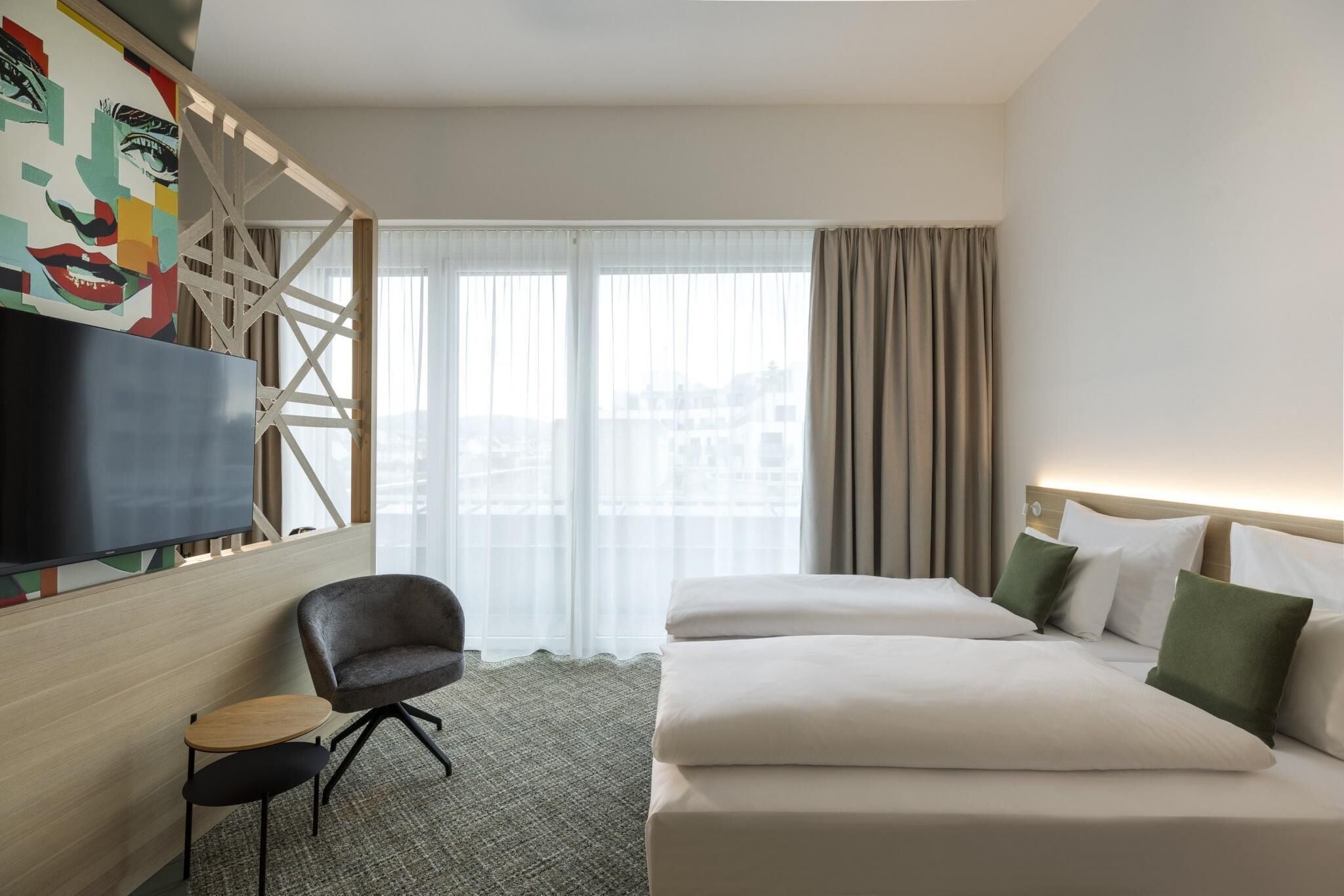 A hotel room with a bed, a chair, a small table, a TV, and a window with curtains.