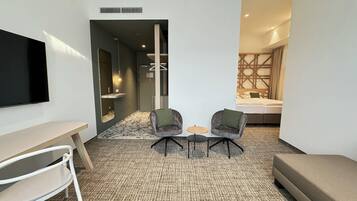 A modern hotel room with a flat-screen TV, a desk, two armchairs, a small round table, and a bed with a geometric-patterned headboard.